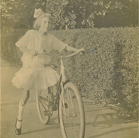 Eudora Welty as a young girl in Jackson, Mississippi, around 1916, riding a bicycle on a hedge-lined sidewalk in a lacy dress, a hair bow, and Mary-Jane shoes.