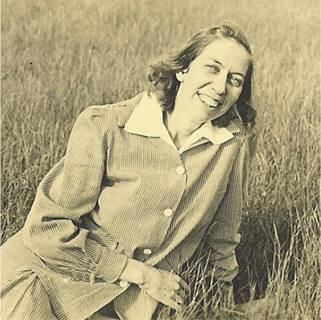 Eudora Welty in her mid-twenties sitting in a grassy field and smiling in a 1930s pin-striped skirt suit and white collared blouse.