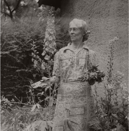 Black-and-white photo of Chestina Welty in the cutting garden behind the stucco garage with snips in one hand and cut flowers in the other.