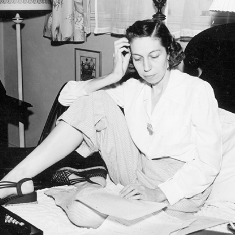 Author Eudora Welty sitting on her bed in a blouse and striped slacks, surrounded by papers and a typewriter, reading a manuscript, with one hand resting on her head.