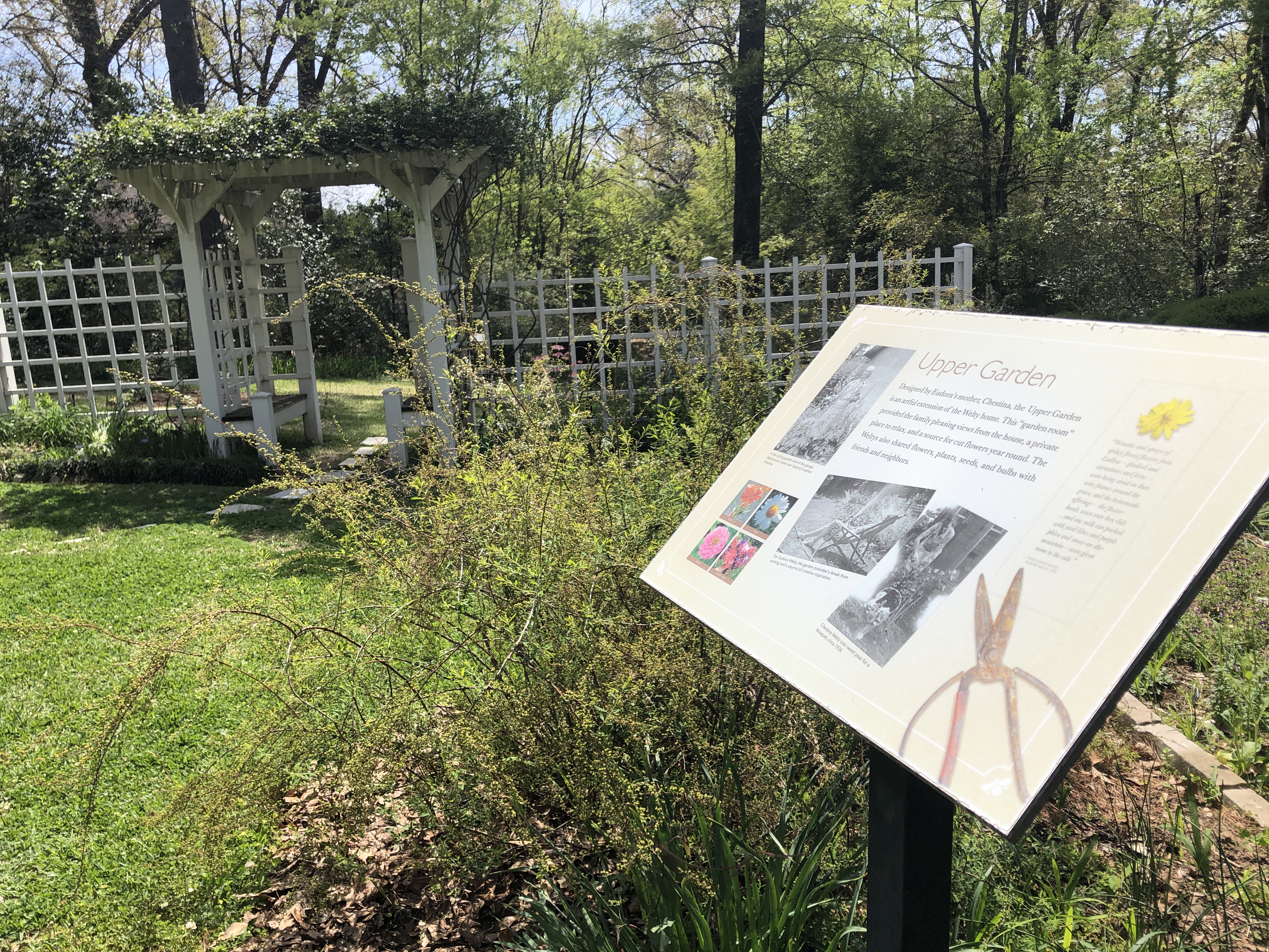 Welty garden interpretive sign in foreground with green Welty lawn and white rose-covered arbor in background
