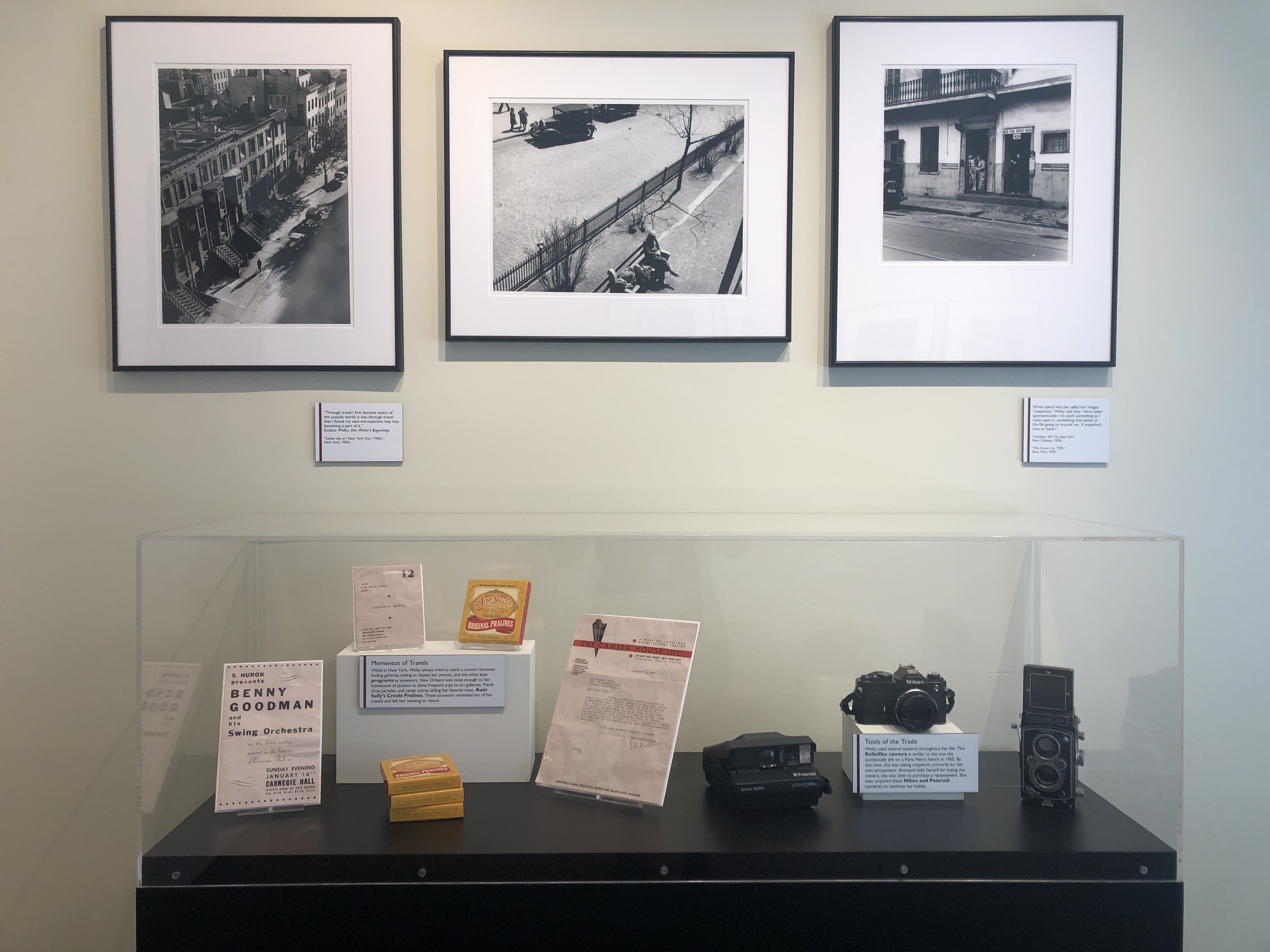 Three black-and-white photographs by Eudora Welty hang in black frames above an acrylic case displaying cameras and photography-related artifacts from the Eudora Welty: Other Places exhibit