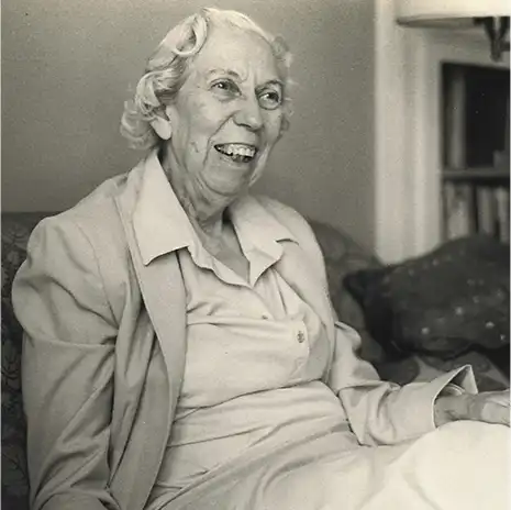 Author Eudora Welty in her later years, seated indoors and laughing, in a dress and blazer.