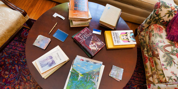 Overhead view of stacks of books on a round, dark wood coffee table between two mismatched floral chairs on a burgundy rug in the Eudora Welty House.