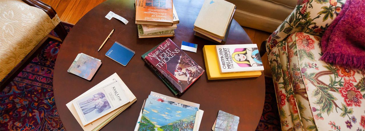 Overhead view of stacks of books on a round, dark wood coffee table between two mismatched floral chairs on a burgundy rug in the Eudora Welty House.